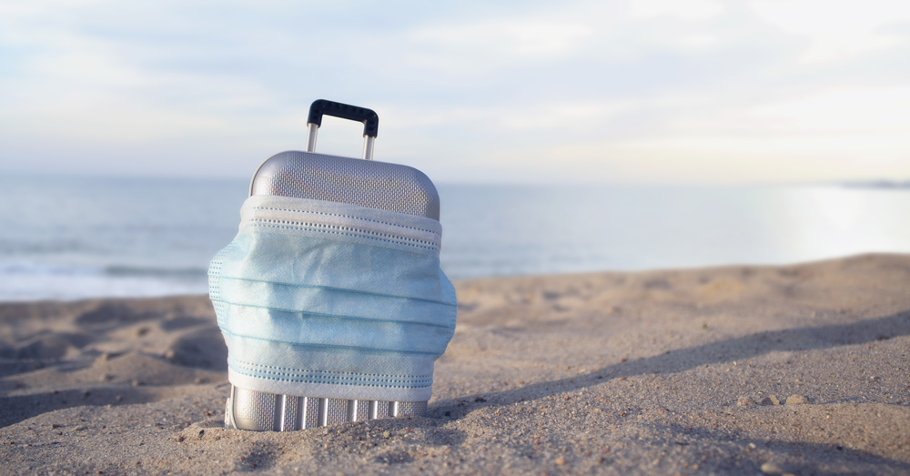 Small suitcase covered by a face mask in the sand by the ocean